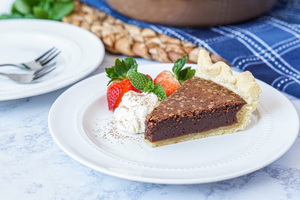 Slice of Chocolate Fudge Pie on a white plate next to whipped cream and two strawberries.