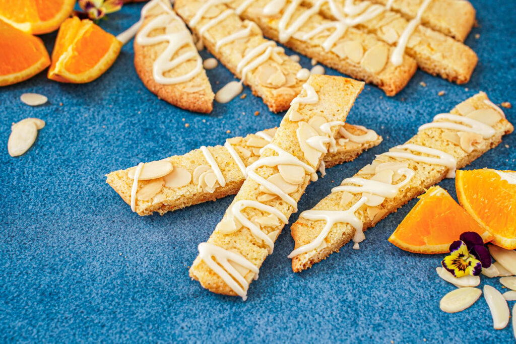 Three Orange Almond Cookie Bars on a blue surface with more bars in the background next to orange wedges and almond slices.