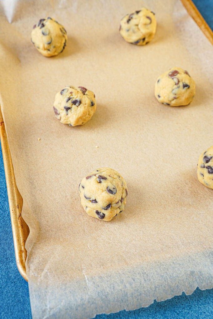 Six balls of chocolate chip cookie dough on a baking sheet.
