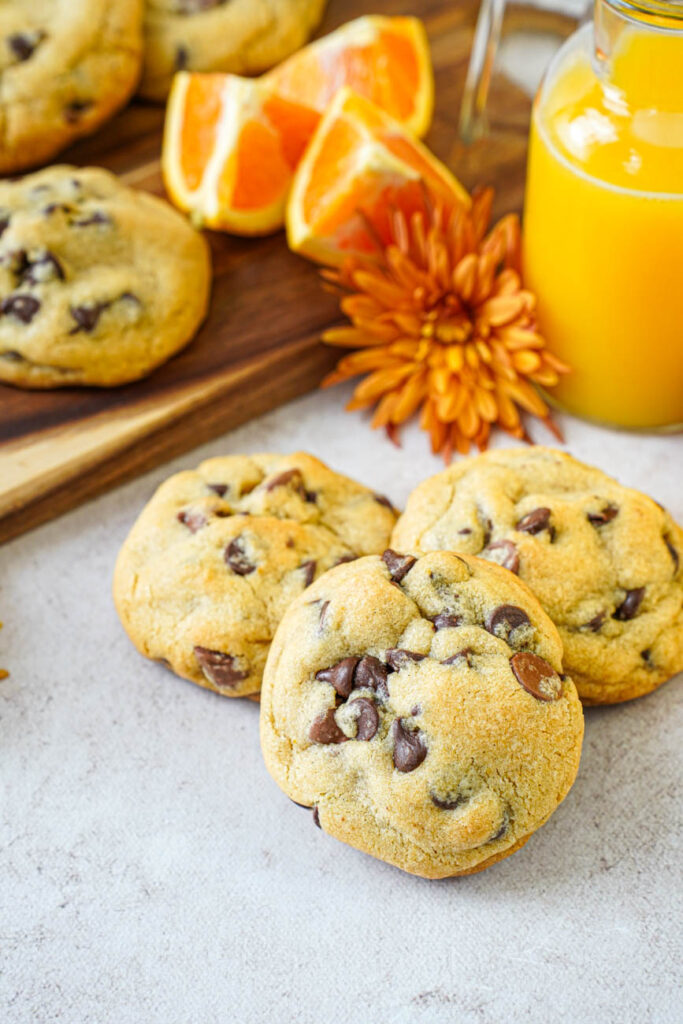 Side view of three Orange Chocolate Chip Cookies with orange juice, orange slices, and orange flowers in the background.