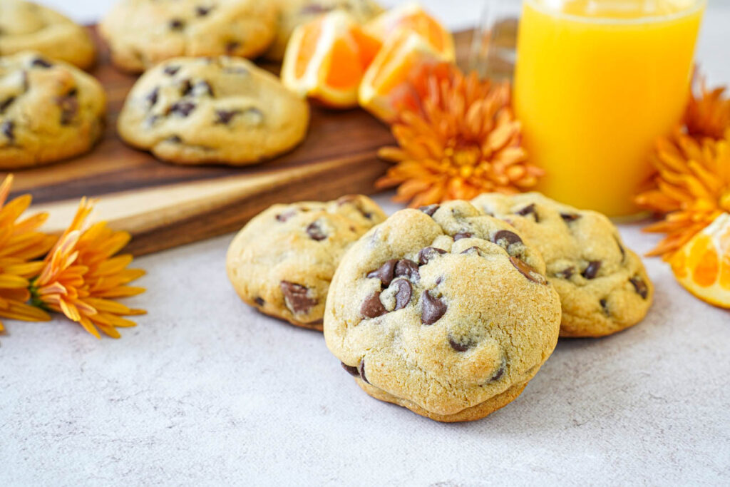 Three Orange Chocolate Chip Cookies with more cookies in the background next to orange flowers and a bottle of orange juice.