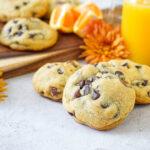Three Orange Chocolate Chip Cookies with more cookies, orange flowers, and a bottle of orange juice in the background.