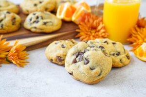 Three Orange Chocolate Chip Cookies with more cookies, orange flowers, and a bottle of orange juice in the background.