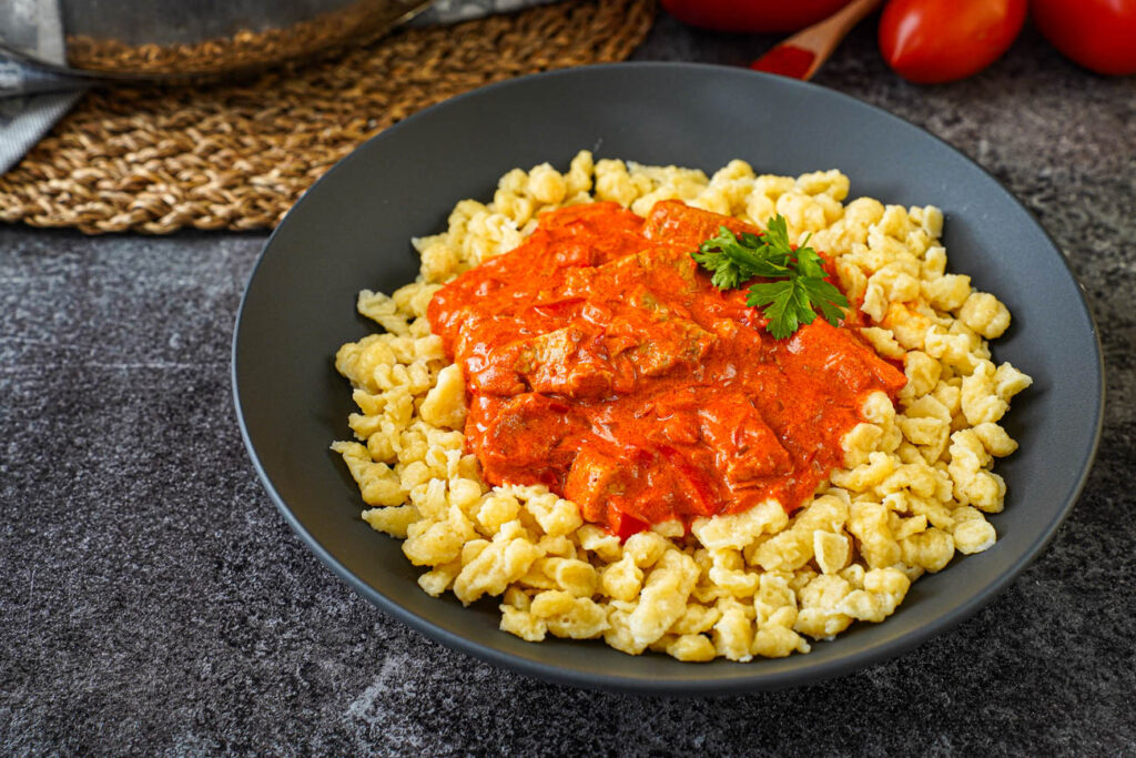 Sertéspaprikás (Hungarian Pork Paprikash) in a black bowl with pasta dumplings and fresh parsley leaves.
