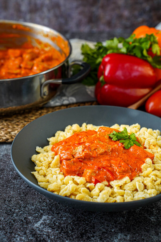Side view of Sertéspaprikás (Hungarian Pork Paprikash) in a black bowl with a pan, bell peppers, and parsley in the background.