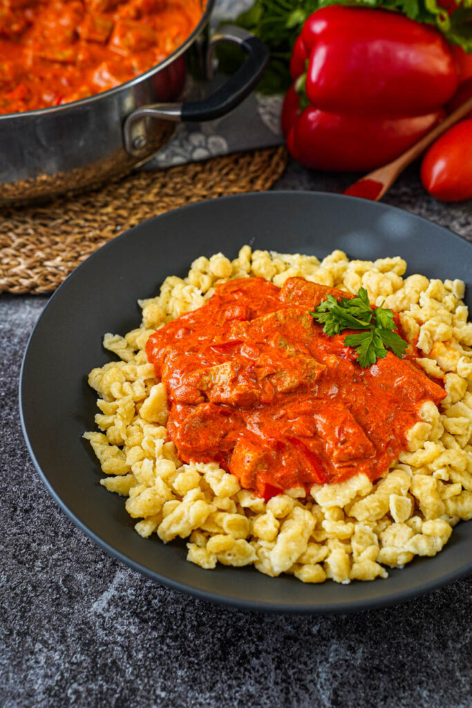 Close up of Sertéspaprikás (Hungarian Pork Paprikash) in a black bowl with more in the background in a pan.