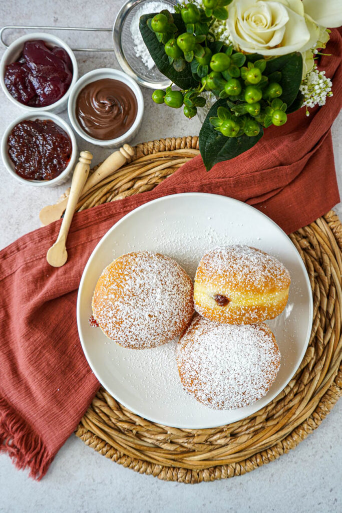 Aerial view of three Berliner (German Filled Doughnuts) on a plate next to jam and white roses.
