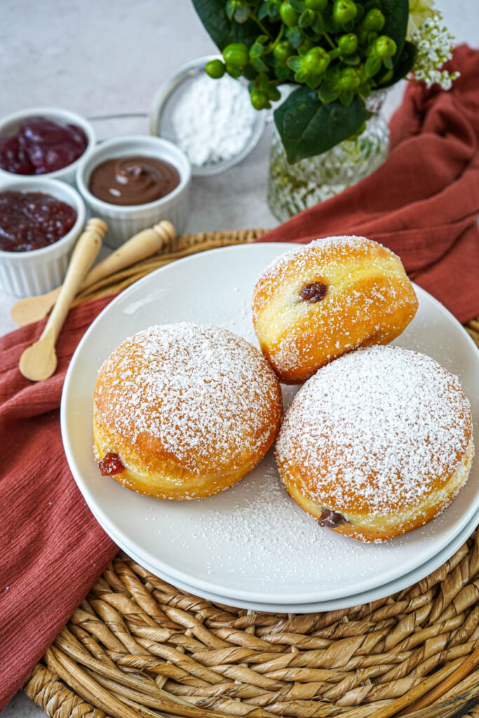 Three Berliner (German Filled Doughnuts) on a white plate with a vase of green leaves and white flowers in the background.