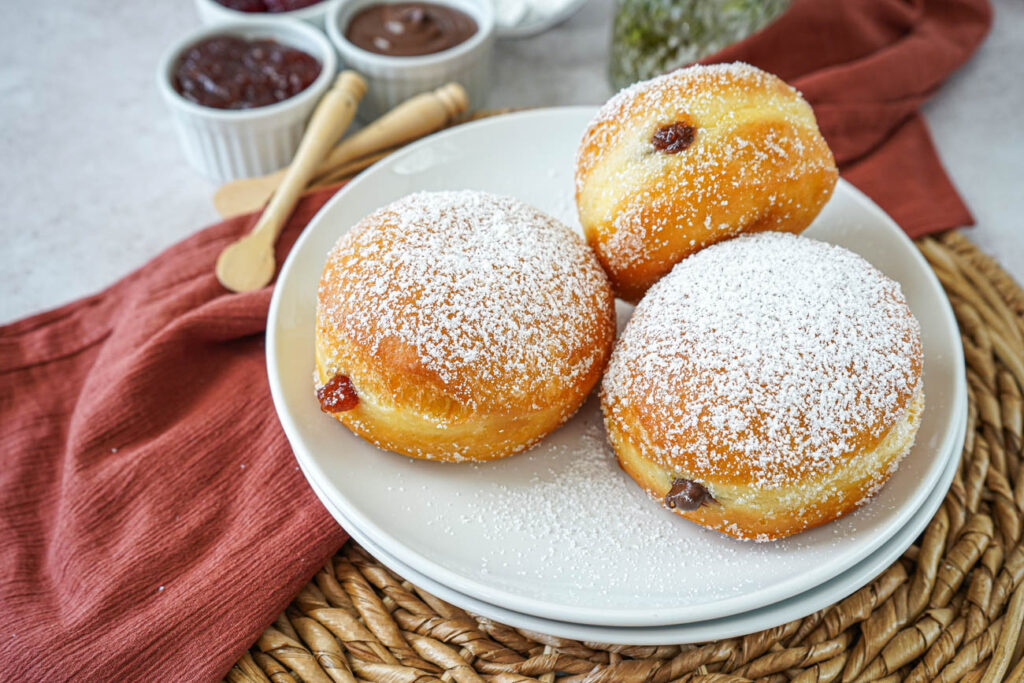 Three Berliner (German Filled Doughnuts) on a white plate next to bowls of red jam and chocolate hazelnut spread.
