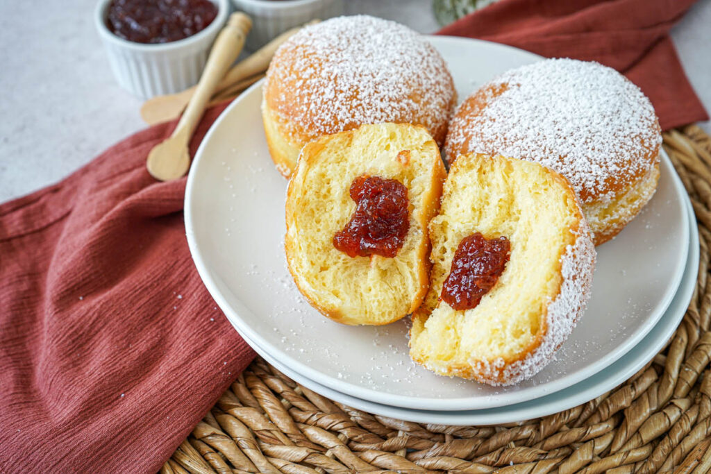 Three Berliner (German Filled Doughnuts) on a plate with one cut in half to show red jam center.