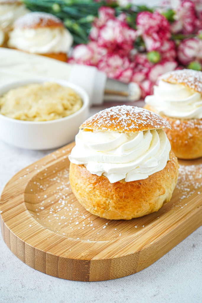 Semlor (Swedish Cream Buns) on a wooden board with almond paste and flowers in the background.