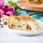 Chocolate Chip Ricotta Pie slice on a white plate with whipped cream next to chocolate chips, pink tulips, and chocolate bunnies.