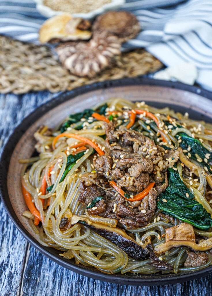Close up of Japchae (Korean Stir-Fried Sweet Potato Noodles) with beef, mushrooms, spinach, carrots, and sesame seeds.