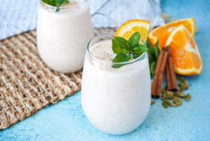 Spiced Orange Lassi in two glasses next to oranges, cinnamon, and cardamom.