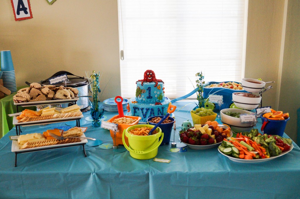 Food at Ocean-themed first birthday with blue tablecloth, snacks in buckets, and white platters.