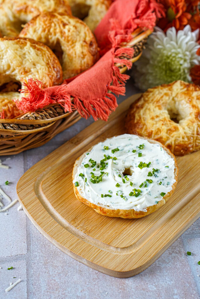 Asiago Bagels in a basket with one on a wooden board and covered in a chive cream cheese spread.