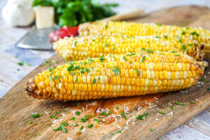 Three Parmesan Garlic Grilled Corn on a wooden board with grated parmesan and chopped parsley.