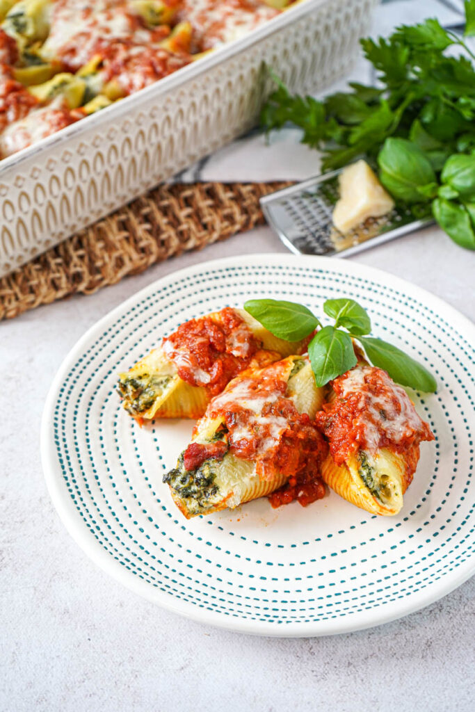 Three Kale and Ricotta Stuffed Shells on a plate with more in a baking dish next to cheese, basil, and parsley.