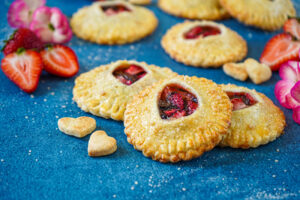 Strawberry Chocolate Hazelnut Hand Pies on a blue surface next to strawberries and pink flowers.