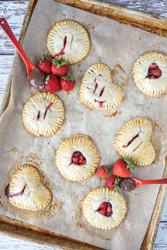 Round and heart Strawberry Nutella Hand Pies on a silver baking sheet with two red spoons holding Nutella next to strawberries.
