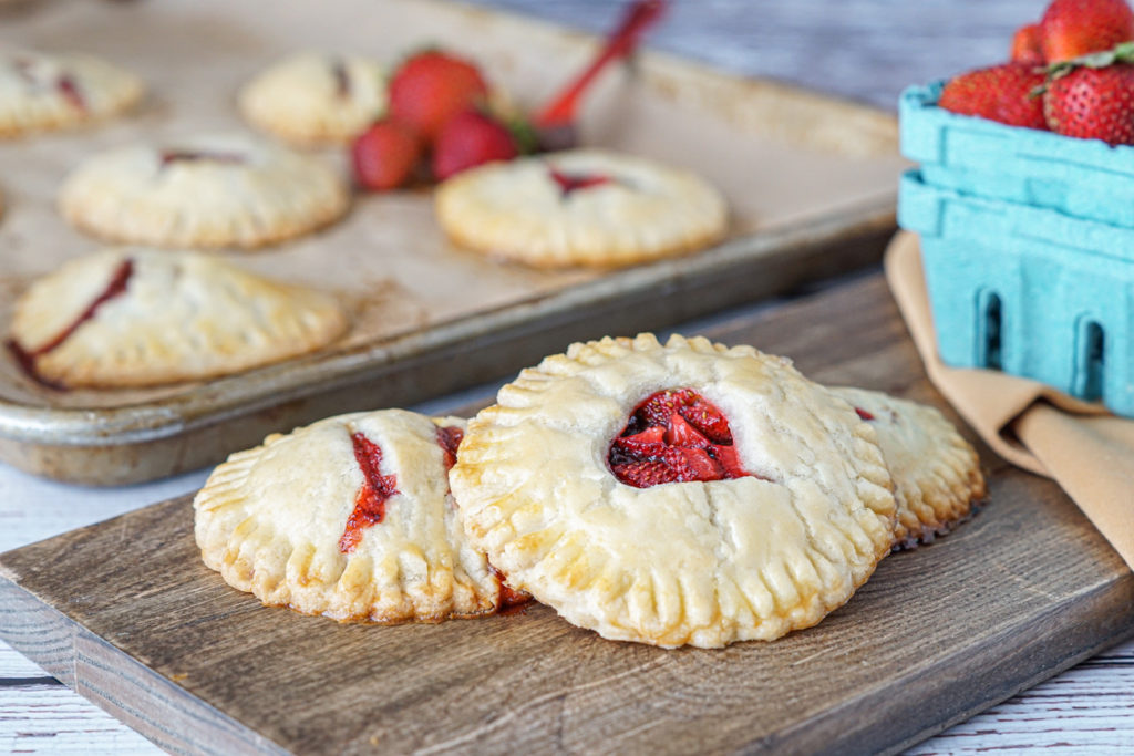 Three Strawberry Nutella Hand Pies on a wooden board next to a blue container of strawberries.
