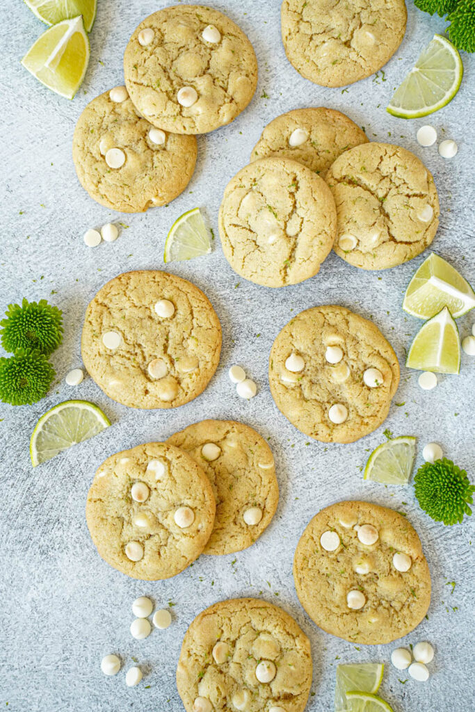 Aerial view of White Chocolate Lime Cookies next to white chocolate chips, lime slices, and green flowers.