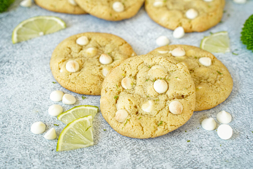 Three White Chocolate Lime Cookies next to white chocolate chips and lime slices.