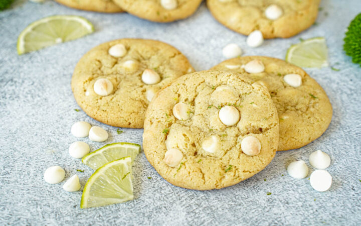 Three White Chocolate Lime Cookies next to white chocolate chips and lime slices.
