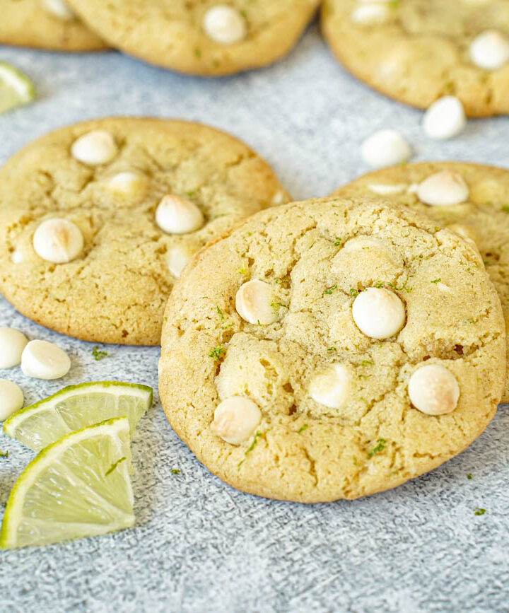 Three White Chocolate Lime Cookies next to white chocolate chips and lime slices.