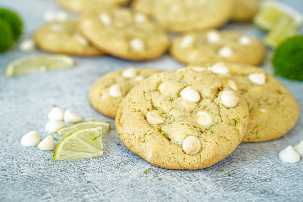 Side view of White Chocolate Lime Cookies next to white chocolate chips, lime slices, and green flowers.