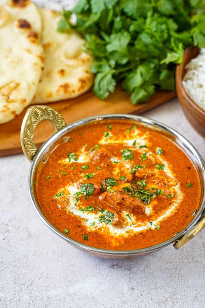 Close up of Murgh Makhani (Butter Chicken) in a bowl with naan, cilantro, and rice in the background.
