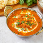 Murgh Makhani (Butter Chicken) in a bowl next to Naan, rice, and cilantro.