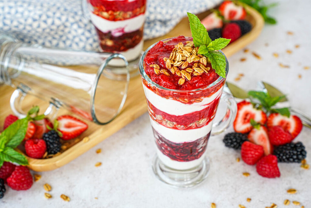 Triple Berry Parfaits in two glasses next to strawberries, blackberries, raspberries, and mint sprigs.