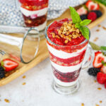 Triple Berry Parfaits in two glasses next to strawberries, blackberries, raspberries, and mint sprigs.