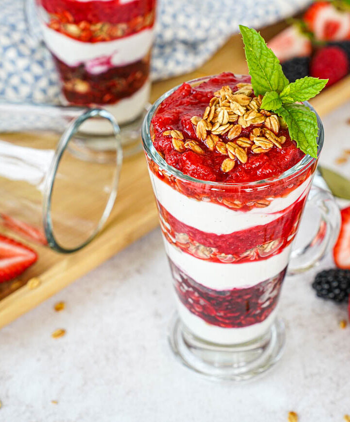 Triple Berry Parfaits in two glasses next to strawberries, blackberries, raspberries, and mint sprigs.