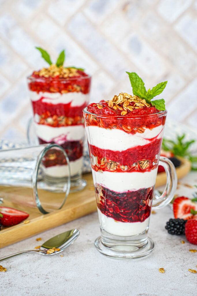 Side view of Triple Berry Parfaits in two glasses next to long spoons, strawberries, raspberries, and blackberries.