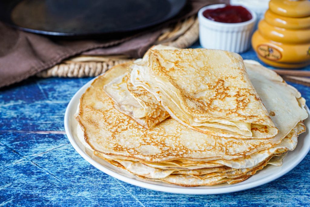 A stack of Blini (Russian Crepes) on a white plate with strawberry jam and honey in background.