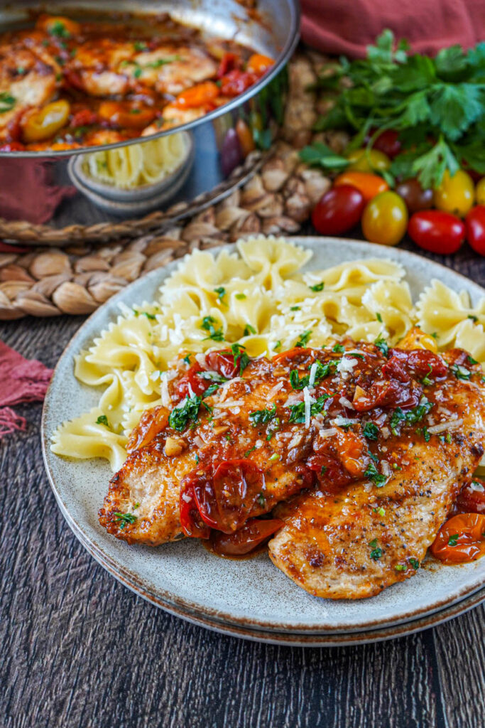 One-Pan Chicken with Cherry Tomatoes on a plate with farfalle pasta with a pan, grape tomatoes, and parsley in the background.