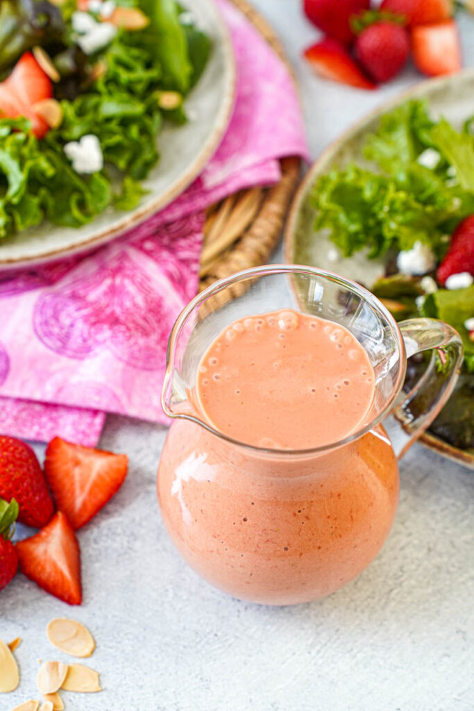 Close up of Strawberry Balsamic Vinaigrette in a glass jar next to plates of salad and fresh strawberries.