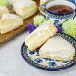 Two Vanilla Cream Cheese Scones on a plate next to a cup of tea and more scones on a wooden board.