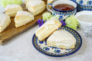 Two Vanilla Cream Cheese Scones on a plate next to a cup of tea and more scones on a wooden board.