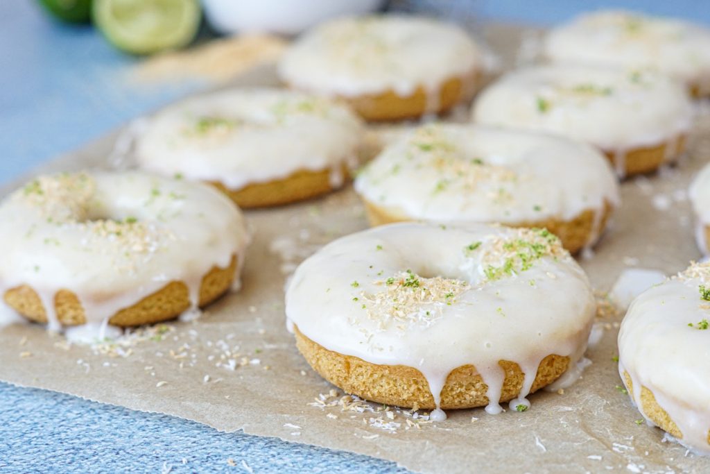 Coconut Lime Baked Donuts on a sheet of parchment paper with lime halves and toasted coconut in the background.