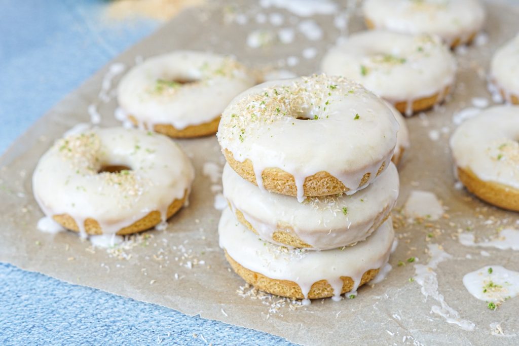 Coconut Lime Baked Donuts on parchment paper with three stacked in the front. 