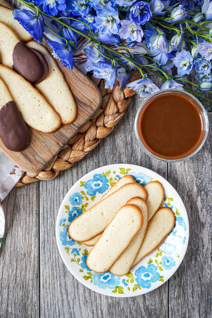 Aerial view of Langues de Chat (French Cat Tongue Cookies) on a plate and wooden board next to a cup of coffee and blue flowers.