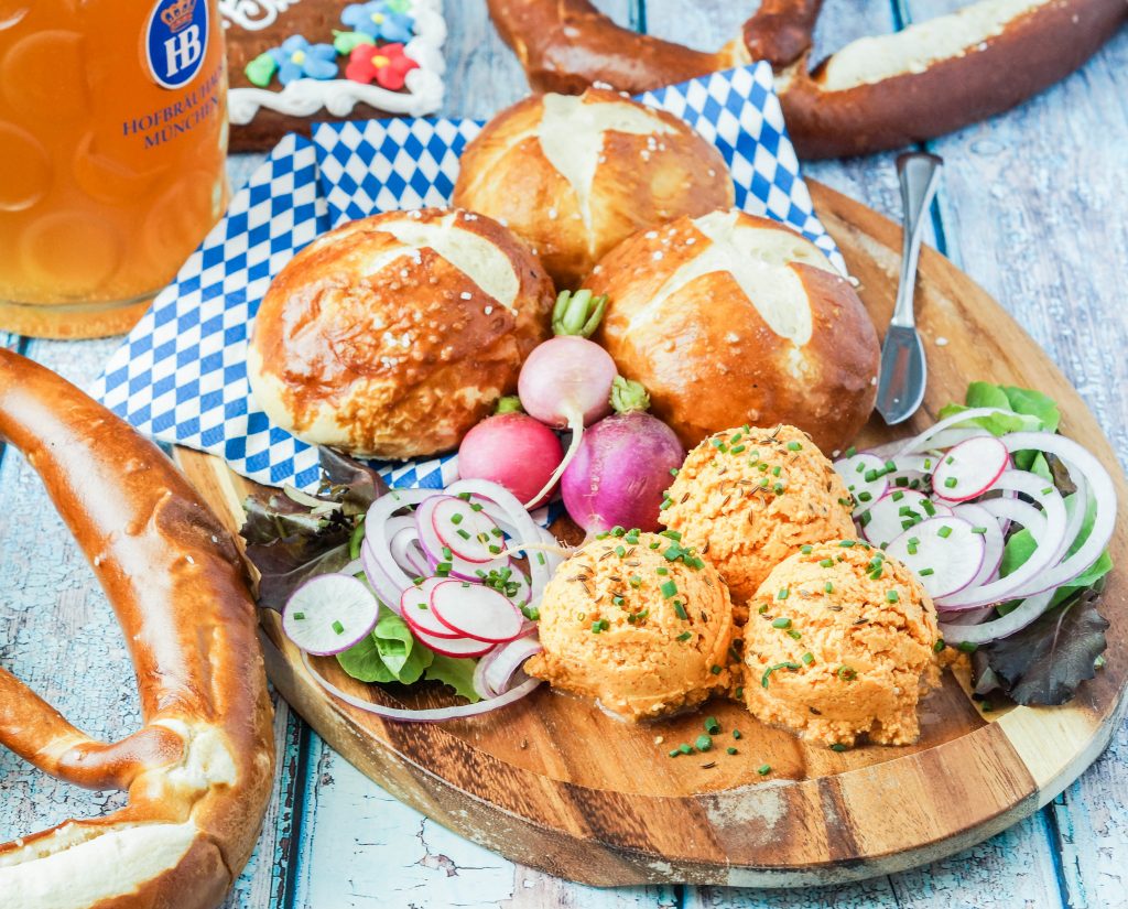 Three scoops of Obatzda (Bavarian Cheese and Beer Spread) on a wooden platter next to pretzels and beer.