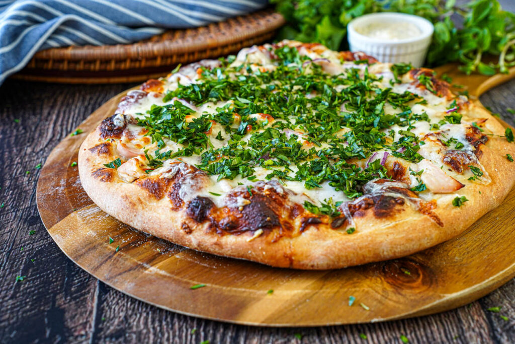 Roasted Garlic Chicken Pizza on a wooden board with fresh herbs and a bowl of garlic sauce in the background.