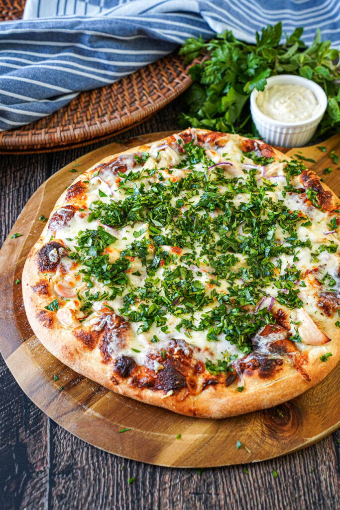 Roasted Garlic Chicken Pizza on a wooden board next to fresh herbs and a bowl of garlic sauce.