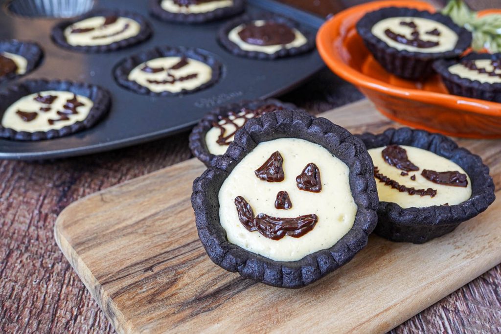 Cheesecake Tartlets with Chocolate Ganache pumpkin faces on a wooden board and in a pan.