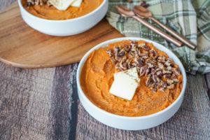 Mashed sweet potatoes in two white bowls with butter and toasted pecans.
