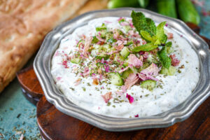 Mast-o-Khiar (Persian Yogurt and Cucumber Dip) in a silver bowl with bread and cucumbers in the background.